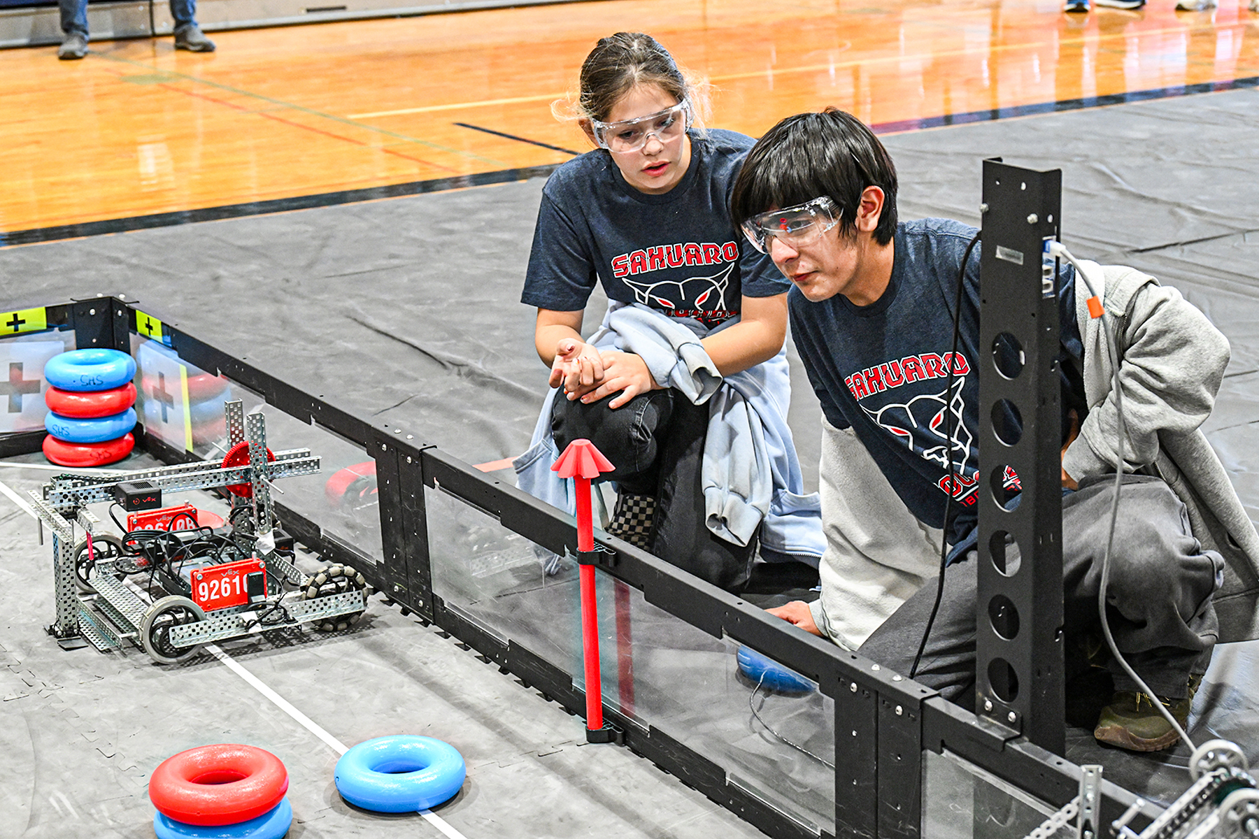 A girl and a boy in safety goggles and Sahuaro shirts look at their robot