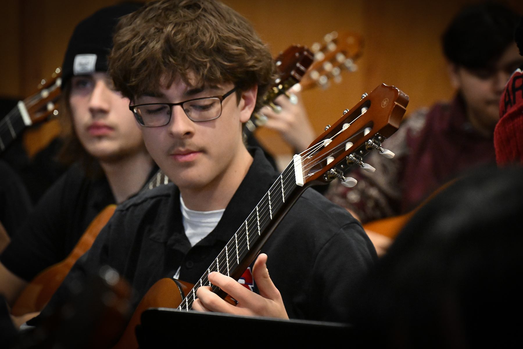 A teen boy with dark hair and glasses focuses as he plays guitar