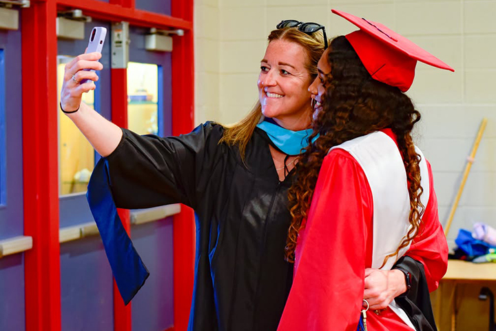A teen girl in a red cap and gown takes a selfie with a teacher