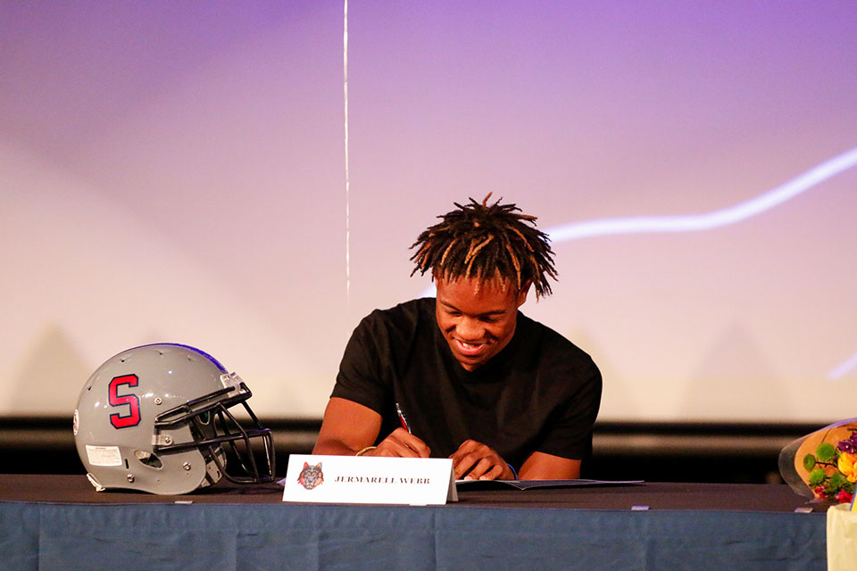 A teen boy signs his letter of intent, with a football helmet on the table in front of him