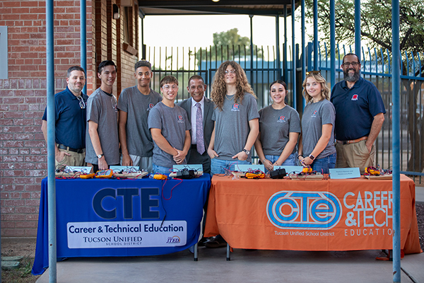 A group of students, their teacher, and the superintendent pose for a photo behind two CTE tables