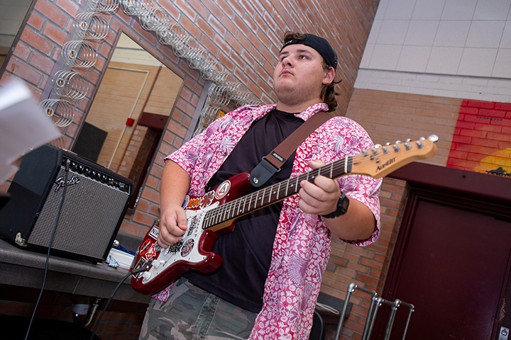 A teen boy in a Hawaiian shirt plays electric guitar