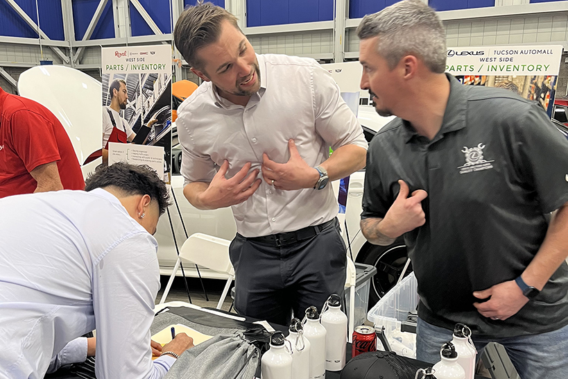 Two men talk while pointing to themselves, while a teen boy leans over to write on a piece of paper