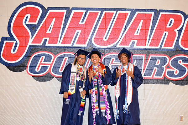 Three teen boys in their caps and gowns pose in front of the Sahuaro mural