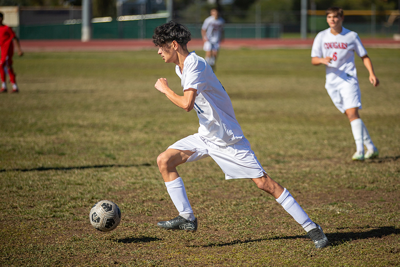 A teen boy in a white jersey and shorts kicks the soccer ball down the field